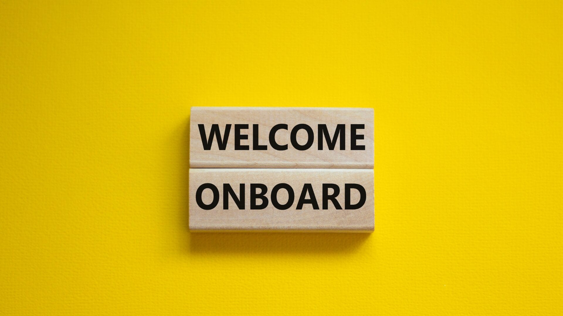 A close-up of two wooden blocks placed side by side on a vibrant yellow background, with the words "WELCOME ONBOARD" written in bold black letters on the blocks