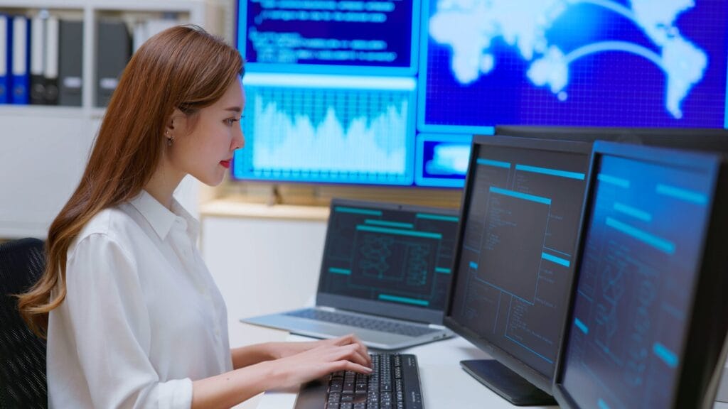 A professional woman in a white blouse works at a desk with multiple computer monitors displaying data visualizations and code interfaces. Behind her, large screens show global network maps and analytics, symbolizing the integration of AI and automation in localization management.