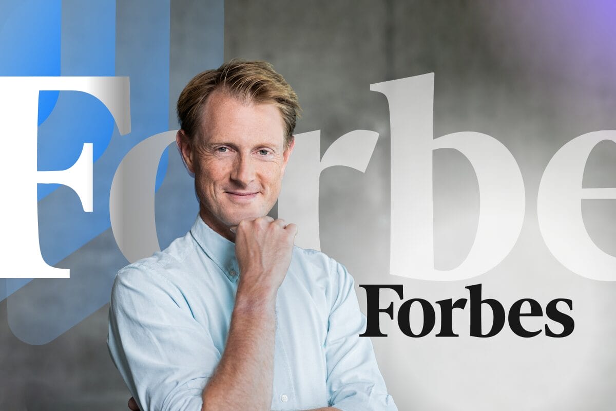 Georg Ell, CEO of Phrase in light blue shirt smiling with hand on chin, posed in front of Forbes logo and Phrase branding elements on gray background