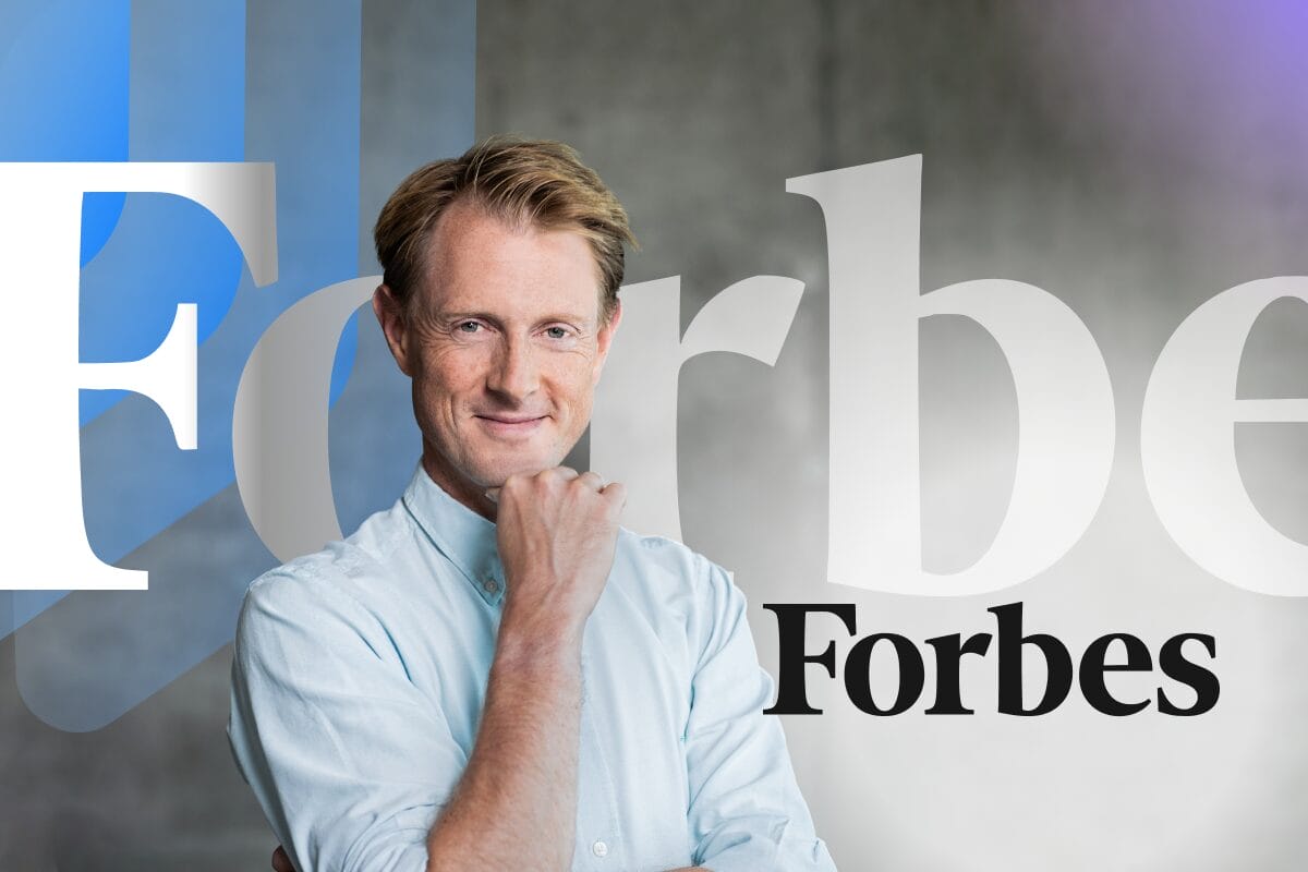 Georg Ell in light blue shirt smiling with hand on chin, posed in front of Forbes logo and Phrase branding elements on grey background